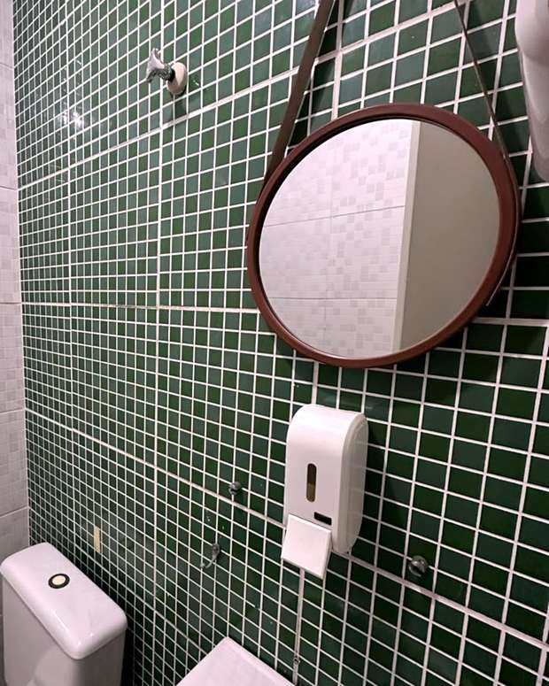 Banheiro da clínica, com parede de pastilhas verdes, espelho redondo e dispenser de sabonete.